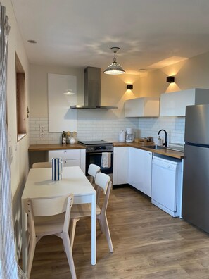 A modern kitchen with white cabinetry, a stainless steel refrigerator, and a dining area with a white table and chairs. - Peaceful cottage overlooking the Rade de Brest, Crozon peninsula Brittany (Roscanvel)