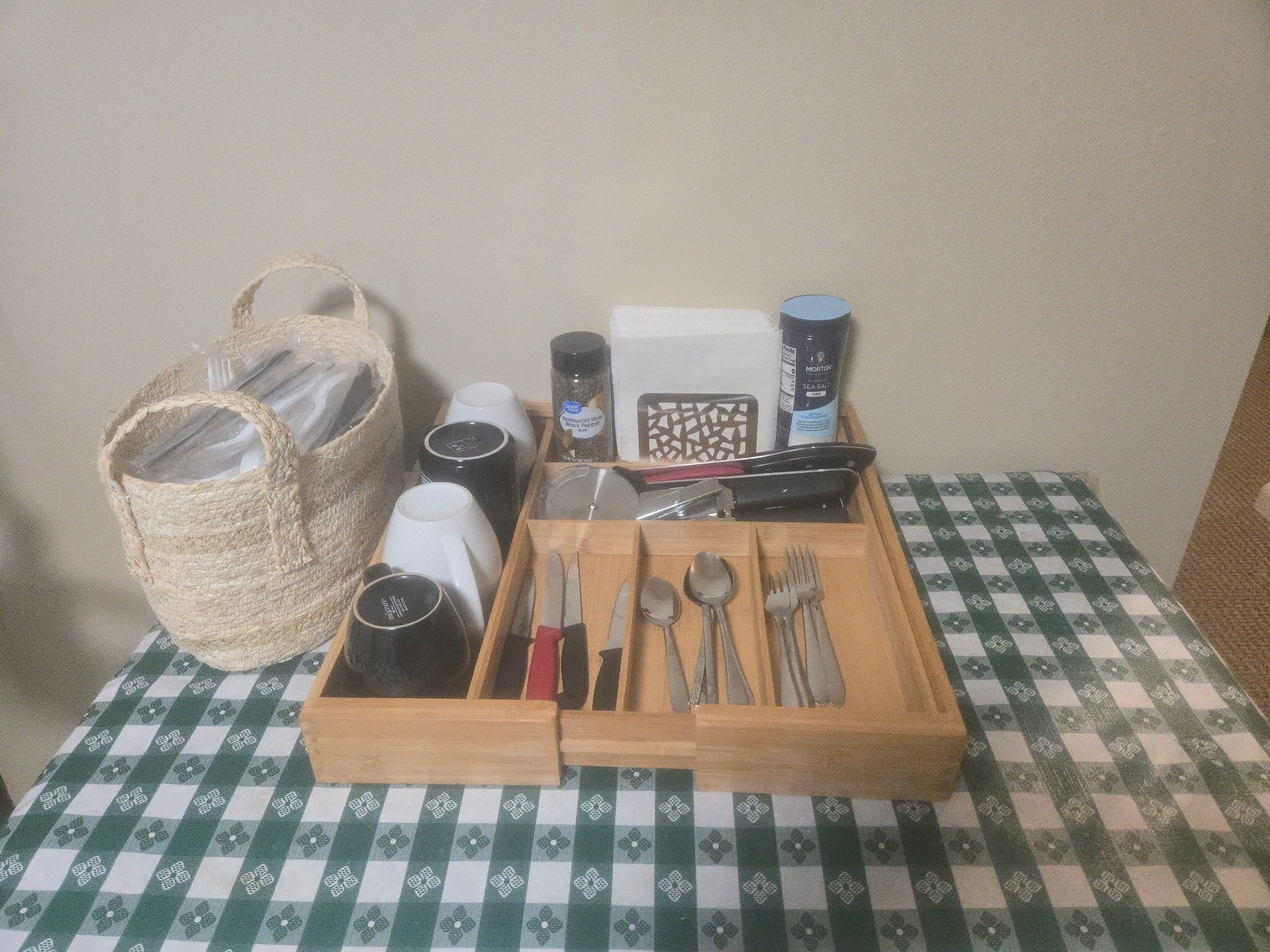 Un plateau en bois avec des couverts, une cafetière, un salier et un poivrier, le tout posé sur une nappe à carreaux.