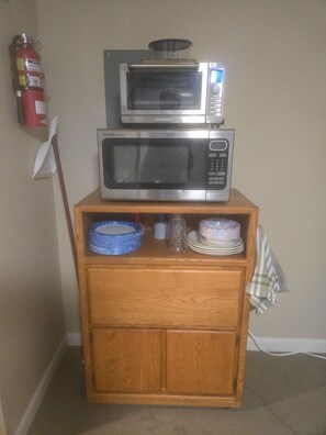A wooden cabinet with two microwaves stacked on top, a fire extinguisher on the wall, and a blue bowl on the lower shelf.