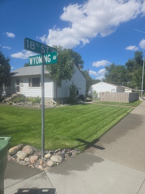 A residential street corner with a house, a lawn, and a clear sky.