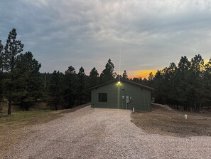 A gravel driveway leading to a small, single-story building with a sloped roof, surrounded by a forested area. - Private, quiet cabin in the trees with farm experience! (Rapid City)