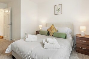 A hotel room with a bed, two bedside tables, a nightstand, a door, and a framed picture on the wall. - Stylish 2-Bed Flat, Leyland – Near Worden Park (Lancashire)