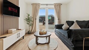 A modern living room with a flat-screen TV, a round coffee table, a black sofa, and a view of a building outside. - Stylish 2-Bed Flat, Leyland – Near Worden Park (Lancashire)