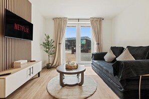 A modern living room with a flat-screen TV, a round coffee table, a black sofa, and a view of a building outside. - Stylish 2-Bed Flat, Leyland – Near Worden Park (Lancashire)