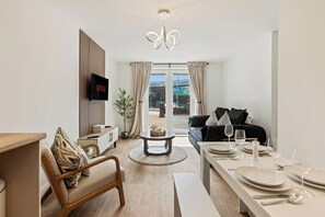 En moderne stue med fjernsyn, sofa, spisebord dækket til to personer og et stort vindue med gardiner.