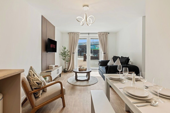 A modern living room with a television, a sofa, a dining table set for two, and a large window with curtains.