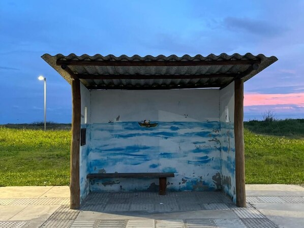 A small, open shelter with a corrugated metal roof, a bench inside, and a mural on the wall depicting a boat on water.
