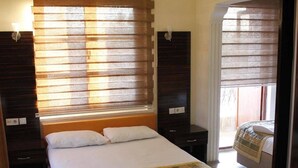 Habitación de hotel con una cama grande, una mesita de noche, una silla y una ventana con persianas.