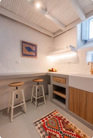 A kitchen with white cabinets, wooden stools, a patterned rug, and a fish-themed artwork on the wall. - 18th-Century Heritage Stone House with Private Garden - Mykonos (Ano Mera)