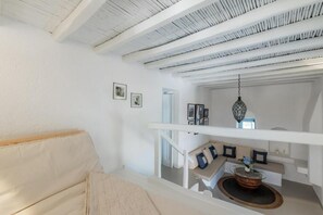 A white interior with a staircase leading to a balcony, a white sofa, and a hanging light fixture. - 18th-Century Heritage Stone House with Private Garden - Mykonos (Ano Mera)