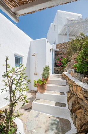 A narrow white-washed street with stone steps leading to a door, surrounded by greenery and a clear blue sky. - 18th-Century Heritage Stone House with Private Garden - Mykonos (Ano Mera)