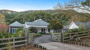 A two-story house with a covered porch, surrounded by a wooden fence and a gravel driveway leading to a gated entrance. - Heronwood - Luxury Country Retreat (Wattamolla)
