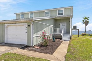 Une maison à deux étages avec un garage, un porche à l’avant et un petit jardin agrémenté de fleurs roses.