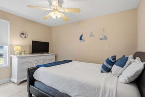 Um quarto com uma cama, uma televisão sobre um penteadeira, um ventilador de teto e detalhes decorativos nas paredes.