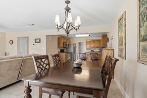A dining room with a wooden table and chairs, a chandelier, and a kitchen in the background.