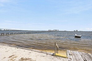 A waterfront view with a wooden pier extending into the water, a sandy beach, and a distant city skyline.