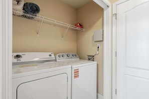 A laundry room with a stacked washer and dryer, a shelf with a blanket, and a wall-mounted electrical outlet.