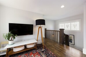 Een moderne woonkamer met een flatscreen televisie, een vloerlamp, een kamerplant, een marmeren salontafel, een tapijtbegroeiing en een trap met een leuning.