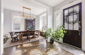 Uma sala de jantar com mesa e cadeiras de madeira, um candelabro e uma janela grande com persianas.