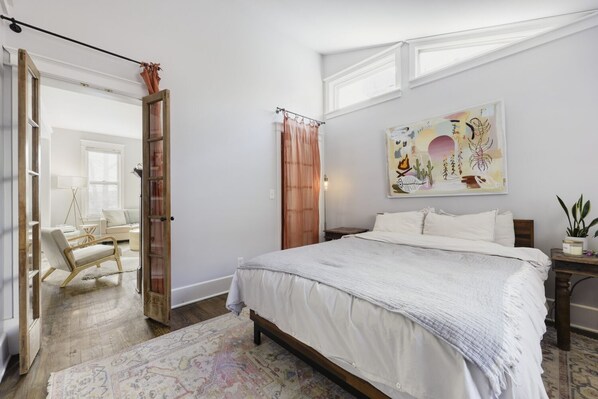 Quarto com uma cama grande, piso de madeira, tapete estampado, um quadro na parede e vista para outro cômodo.