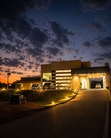 Un edificio moderno con entrada cubierta, varios autos estacionados al frente y un camino de acceso bien iluminado.