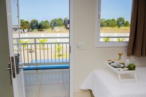Una habitación con balcón, una cama con mesita y una ventana con vistas a árboles y una valla.