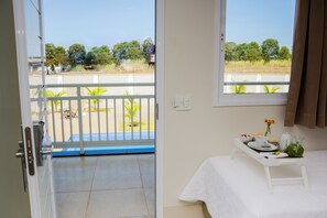 Una habitación con balcón, una cama con mesita y una ventana con vistas a árboles y una valla.