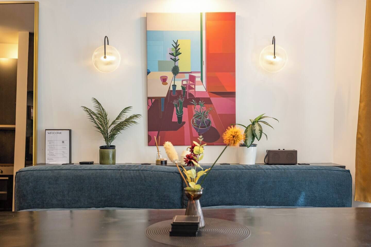 Een receptiegedeelte met een blauwe bank, een bord met een welkomstboodschap, een schilderij en twee wandlampen.