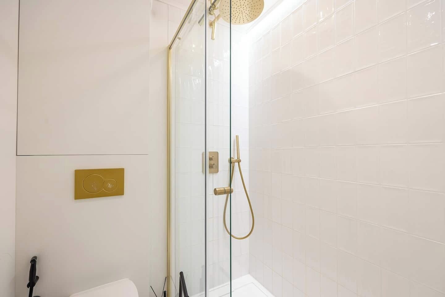 Een moderne badkamer met een douchecabine van glas, een gouden douchekop en een witte betegelde wand.