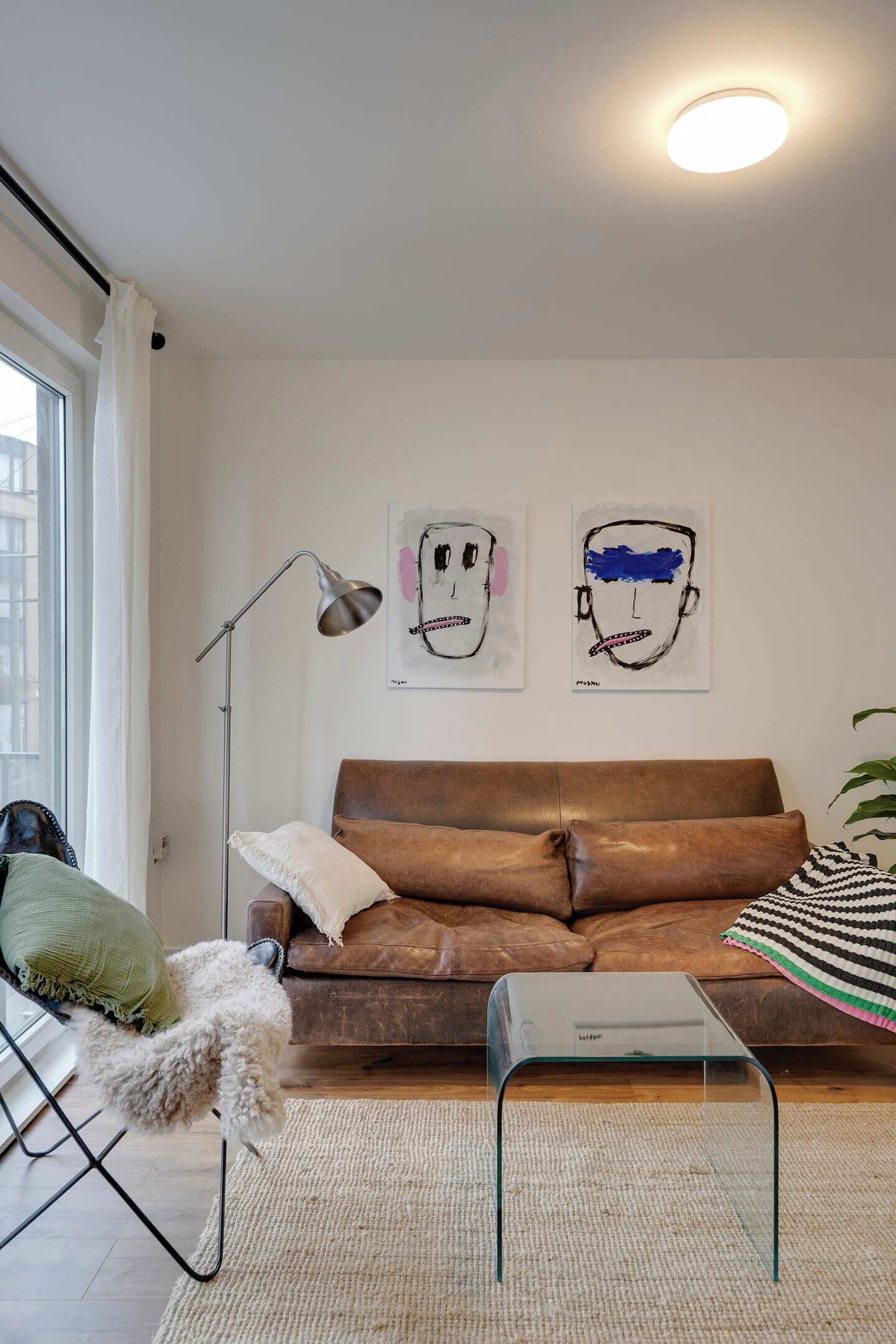 Uma sala de estar com um sofá de couro marrom, uma mesa de centro de vidro, um tapete branco, um abajur e duas pinturas abstratas na parede.