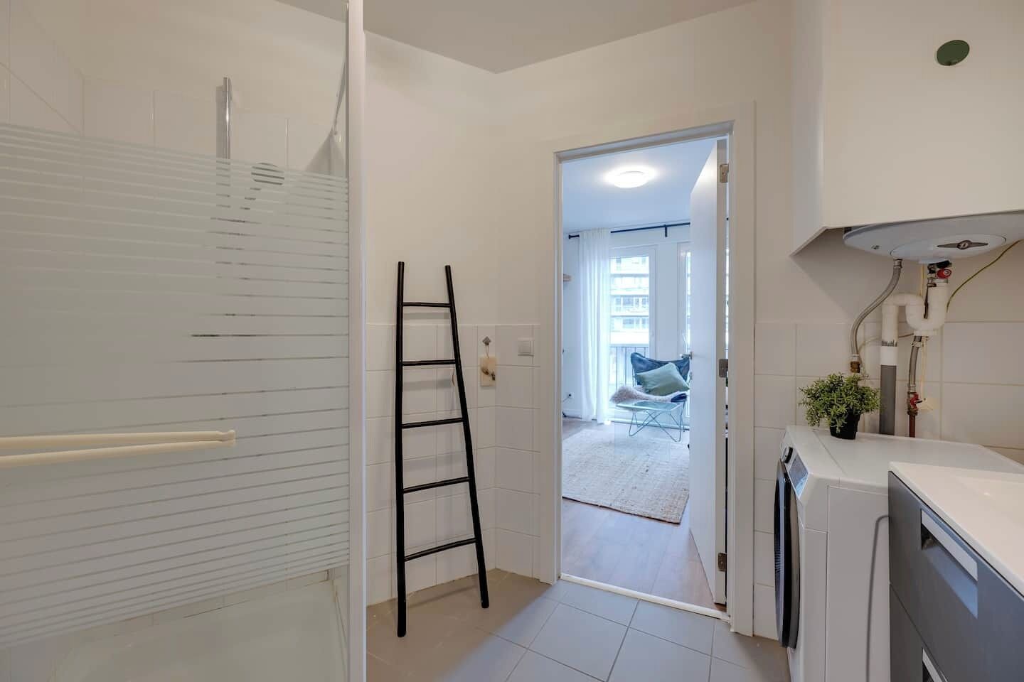 Een moderne badkamer met een douche, een ladder en uitzicht op een woonruimte met een stoel en een plant.