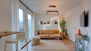 Een moderne woonkamer met een bank, een salontafel, een eethoek met een stoel en een aan de muur bevestigde televisie.