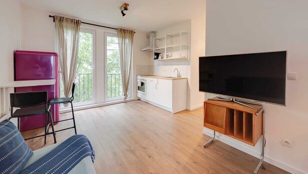 Ett modernt vardagsrum med en platt-TV, en soffa, en matplats med kök och en balkong med gardiner.
