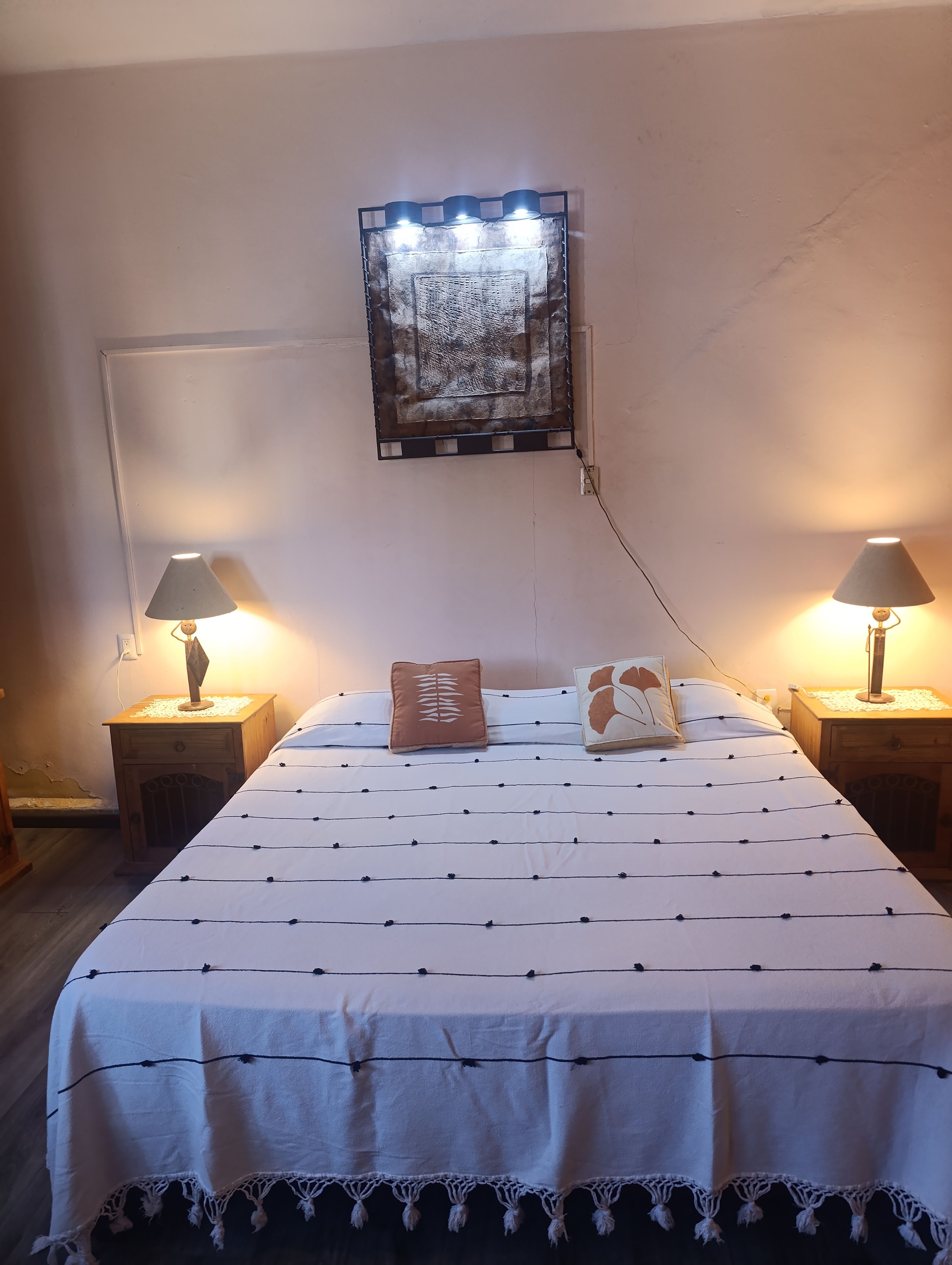A neatly made bed with a patterned bedspread, two bedside lamps, a wall-mounted light fixture, and a small wooden nightstand with a lamp.