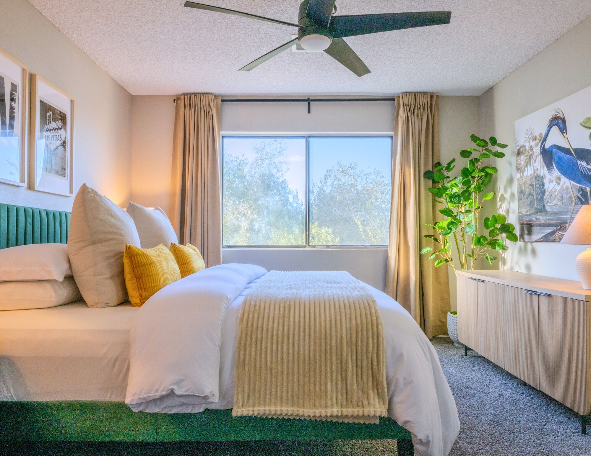 Una habitación de hotel con cama, ventilador de techo, ventana con cortinas, una planta y obras de arte en las paredes.