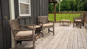 Una terraza de madera con dos sillas, una mesa, un ventanal y vistas a un paisaje verde.