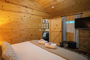 Ein Zimmer in einer Holzhütte mit Bett, Fernseher, einer Kommode und einem Fenster mit Vorhängen.