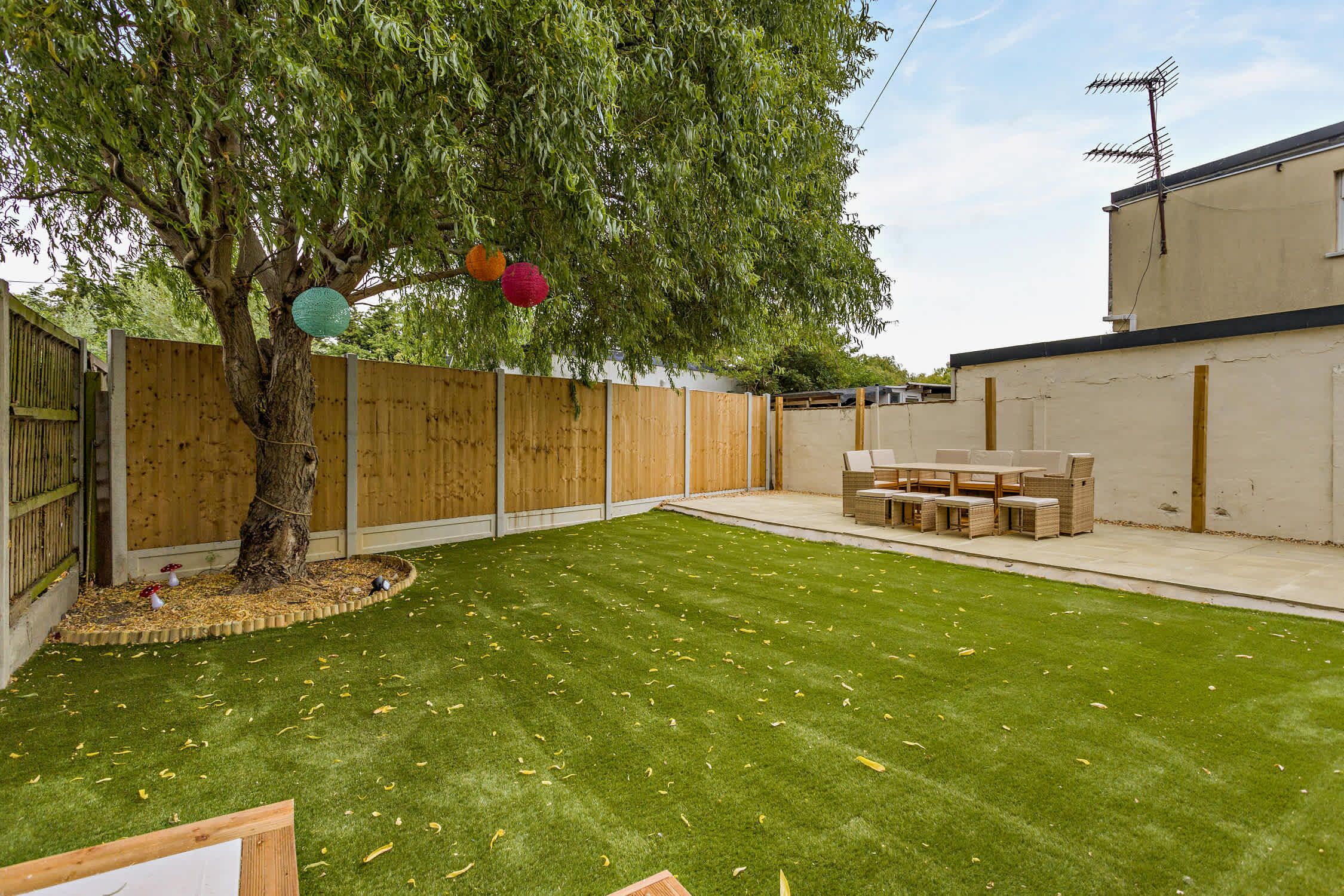 Un jardin avec une clôture en bois, un arbre, une pelouse et une terrasse aménagée avec du mobilier d’extérieur.