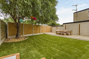 A backyard with a wooden fence, a tree, a grassy lawn, and a patio with outdoor furniture.
