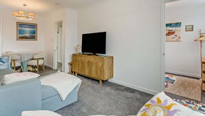 A modern living room with a flat-screen TV on a wooden cabinet, a sofa, a dining table with chairs, and artwork on the walls.