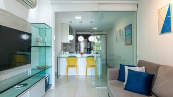 A modern living space with a TV, glass dining table, yellow chairs, a sofa with blue cushions, and a kitchen area with a refrigerator and hanging lights.