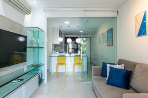 A modern living space with a TV, glass dining table, yellow chairs, a sofa with blue cushions, and a kitchen area with a refrigerator and hanging lights. - Nannai Residence Muro Alto Beira Mar (Ipojuca)