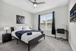 Ein modernes Schlafzimmer mit Bett, Nachttischen, Schreibtisch, Stuhl, Deckenventilator und einem Fenster mit Jalousien.