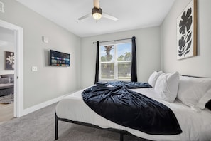 A bedroom with a bed, a ceiling fan, a window with blinds, and a wall-mounted TV.