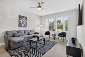 A modern living room with a sectional sofa, a coffee table, a floor lamp, and a ceiling fan. There are two windows with blinds, a painting on the wall, and a small table with a vase. - Awesome 2BR Apt/Pool/Gym/King Bed/Beachwalk access (St. Johns)