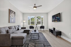 A modern living room with a sectional sofa, a coffee table, a TV mounted on the wall, and a ceiling fan.