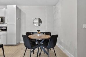A modern kitchen with white cabinetry and a microwave, a dining area with a round wooden table and black chairs, and a round mirror on the wall.
