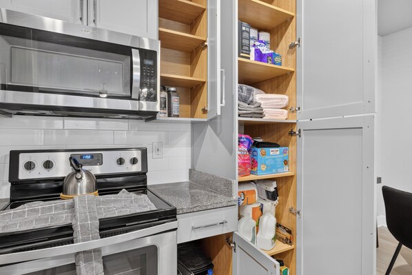 A kitchen with a microwave, stove, and oven. There are shelves with various items including food packages and bottles.