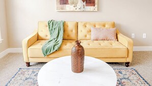 Ein gelbes Sofa mit einer grünen Decke und einem gemusterten Kissen, eine weiße Vase auf einem runden Tisch und ein Wandbild darüber.