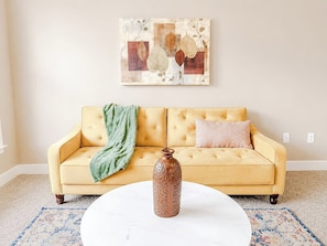A yellow sofa with a green throw blanket and a patterned pillow, a white vase on a round table, and a wall art piece above.